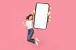 Full length of cheerful Asian woman jumping and smiling in air with showing cellphone blank screen with empty space for mobile app on screen. Isolated in studio pink background. Creative collage.