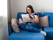 © pijav4uk - Woman with dark hair in a T-shirt sitting on a blue sofa in her living room and reading the news. Female in glasses at home reading article in magazine. Studying at home.