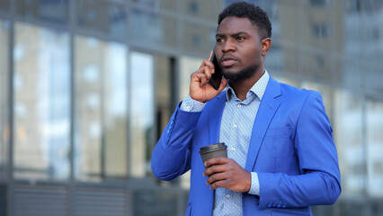 African American man takes break from office chaos holding coffee cup. Stylish businessman talks on phone with friends discussing future weekends