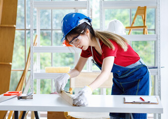 Wall Mural - Asian professional female engineer architect foreman labor worker wears safety goggles glasses using pencil and square angle degree ruler measuring wood plank on working table with laptop computer