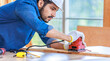 Indian professional bearded male engineer architect foreman labor worker carpenter wears safety helmet and gloves using wood polishing machine grinding sanding wooden board surface on workbench