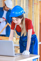 Wall Mural - Asian professional female engineer architect foreman labor worker wears safety goggles and jeans apron standing smiling using laptop computer working on wooden table with tools in construction site