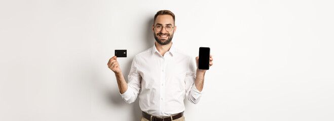 Business and online payment. Smiling handsome man showing mobile screen and credit card, standing over white background