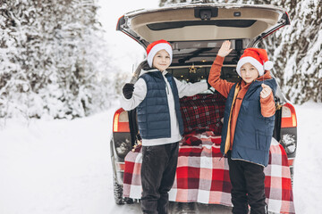 Wall Mural - Two friends teenagers boys in red santa hats with with sparkles Bengal fire standing near trunk of car decorated for Christmas and New Year in snowy winter forest. Road trip and local travel.