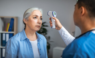 Wall Mural - Adult asian male doctor checking temperature with contactless thermometer to sad senior lady patient