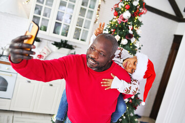 a child in a sweater with deer and in a santa hat sitting on dad's shoulders smiles and communicates with relatives via video link against the backdrop of a christmas tree.