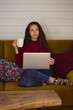 © vita - White brunette girl sits on couch holding white cup of tea, coffee in hand and laptop, looking up dreamily. European young woman shopping online, working remotely, studying at home at autumn or winter