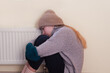 A girl in warm clothes warms herself by the radiator. Energy crisis. Heating problems.