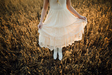 Low Section Of Girl Wearing White Dress Standing Amidst Plants On Field During Sunset