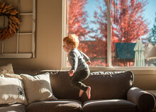 Toddler Boy Jumps On Couch In Living Room In Front Of Fall Window