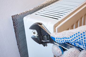 Plumber with wrench repairs a metal heating radiator forming part of a central heating system with energy-efficient thermal insulation on the wall.