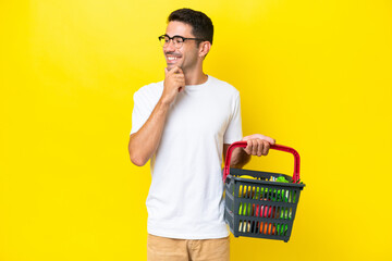 Young handsome man holding a shopping basket full of food over isolated yellow background looking to the side and smiling