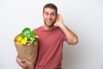 Wall Mural - Young caucasian man holding a grocery shopping bag isolated on white background listening to something by putting hand on the ear