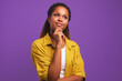 Young pretty ethnic African American woman thinking holding hand on chin imagining happy future or coming up with new creative ideas dressed in stylish casual clothes stands on purple solid background