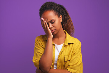 Young tired African American woman closing eyes making face palm gesture after perpetration of serious mistake or experiencing unpleasant emotions standing in purple studio. Depression, stress concept