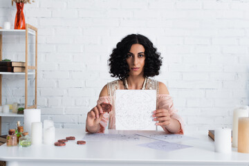 Wall Mural - brunette astrologist showing constellations map and clay rune while looking at camera.