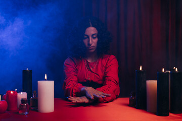 Wall Mural - fortune teller sitting during magic session near burning candles on dark background with blue smoke.