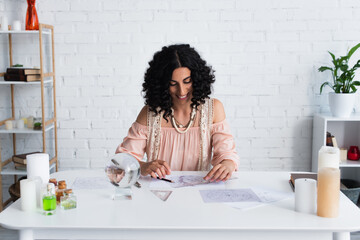 Wall Mural - smiling astrologer looking at celestial charts near crystal ball and bottles with essential oils.