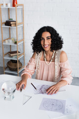 Wall Mural - brunette astrologer smiling at camera near crystal ball and star charts.