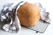 © tilialucida - Home baked farmhouse mixed bread. Close-up on loaf of bread baked in baking machine. Whole wheat bread. Rustic organic farmhouse bio product on drying rack with stripy towel on white table.