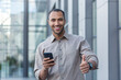 © Liubomir - Young African American businessman outside office building smiling and looking at camera, thumbs up, programmer using smartphone for app testing.