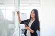 Portrait of young Asian woman writing on whiteboard in office holding smartphone