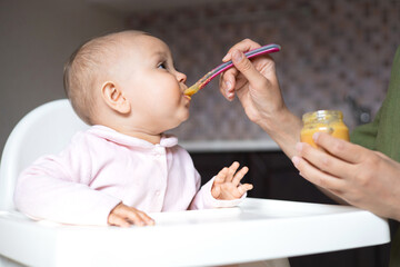 baby food. a baby in a high chair eats vegetable puree from a spoon. mom feeds the baby from a jar. 