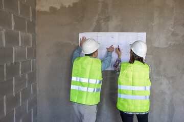 Wall Mural - two engineers, a man and a woman in white helmets and protective vests, are standing in the room and holding a plan of the construction site and fire safety in their hands. interior designers