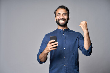 Happy indian business man investor winner using cell mobile phone isolated on gray background. Excited ethnic guy holding smartphone feels surprised about betting win on cellphone, celebrating success