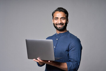 Happy smiling indian business man employee or manager standing isolated on gray background holding laptop advertising online products, business trainings and webinars, websites or services. Copy space