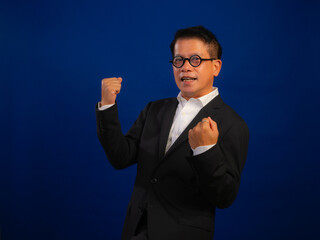 Portrait of happy middle-aged asian confident successful businessman wearing suit celebrating on blue background in studio, looking glad cheerful and positive professional leadership