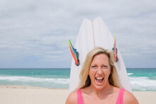 Head Shot Of Woman In Bathers Looking At Camera On Beach Holding Surfboard Behind Head