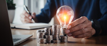 Businessman Holding And Putting Lightbulb On Coins Stack On Table For Saving Energy And Saving Money Concept.