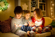 Two Big Sisters And Their Toddler Brother Playing With Chistmas Lights In A Cozy Living Room On Christmas Eve. Kids Spending Time At Home During Winter Break.