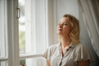 © svetograph - A middle-aged woman in a cream dress sits mysteriously and looks out the window on the windowsill. Green trees outside.