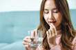 © jumlongch - Sick Asian woman eating pills with a glass of water in hand in her house, close up to a glass of water.