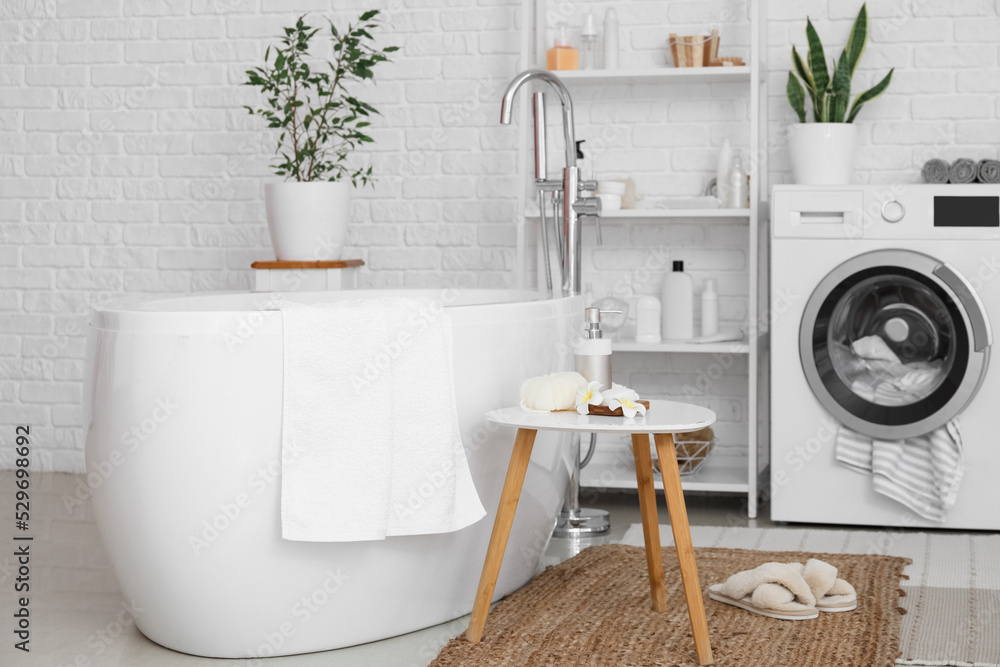 Modern bathtub and table with bath accessories in light room