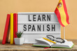 Board with text LEARN SPANISH, flag, eyeglasses and books on table against beige background