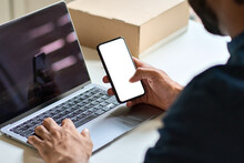Male Hand Holding Smartphone With White Mock Up Screen Using Laptop At Work Desk. Business Man Showing Blank Mobile Display Mockup Template Of Cell Phone With Empty Space, Over Shoulder View.