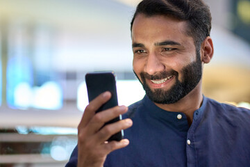 Wall Mural - Smiling happy indian business man, eastern professional businessman holding cellphone tech using mobile phone looking at cell screen watching social media standing in urban city on street outdoors.