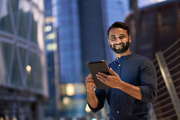 Wall Mural - Smiling happy young eastern indian business man professional manager standing outdoor on street holding using digital tablet online fintech in night city with urban lights looking at camera, portrait.