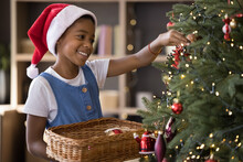 Happy Cute African American Kid In Red Santa Cap Hanging Up Ornament Baubles On Christmas Fir Tree Green Branches, Enjoying December Holiday Time On Xmas Eve, Celebrating New Year. Childhood Concept