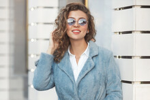 Portrait Of Happy Millennial Woman 30-35 Years Old With Curly Hair In Blue Trendy Sunglasses And Faux Fur Coat And White Shirt