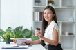 © amnaj - At the office, a young Asian businesswoman sit and relaxe with a cup of coffee and a bright smile on her laptop.