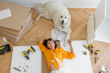 Wall Mural - Young happy woman lies on floor with her cute dog, resting while making repairing at new apartment, top view. Young woman assembling furniture by herself. DIY concept