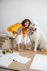 Wall Mural - Young woman makes selfie photo on phone with her cute dog, have fun while making repairing at new apartment. Happy improvement and house repairing concept