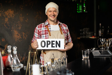 Wall Mural - Smiling male bar owner stands behind the bar and holds the sign open. He invites everyone to enter. Open.