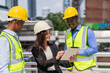 © Chaiya - caucasian businesswoman in black suit wear helmet working on tablet at construction site outside building in city and standing near black engineer colleagues