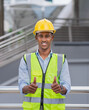 portrait of black engineer in glow shirt, smile, wear helmet, give thumb up, looking at camera at construction site outside building in city