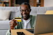 Happy middle aged african american guy in glasses and casual enjoys online shopping on laptop with credit card